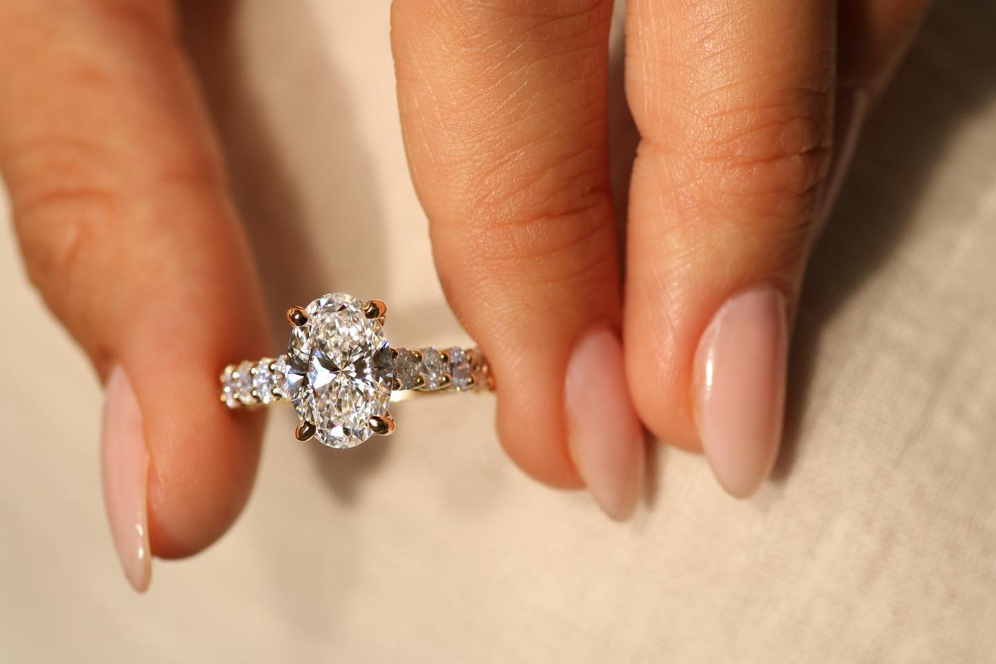 Gold ring with an oval-cut diamond center stone and diamond-encrusted band held between manicured fingers.