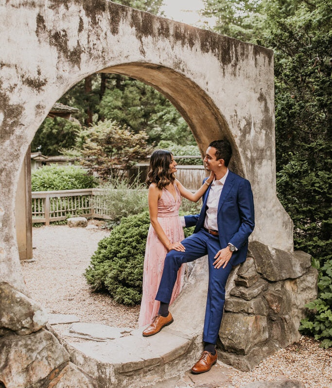 Couple posing in a stone archway; woman in pink dress, man in blue suit with brown leather shoes.