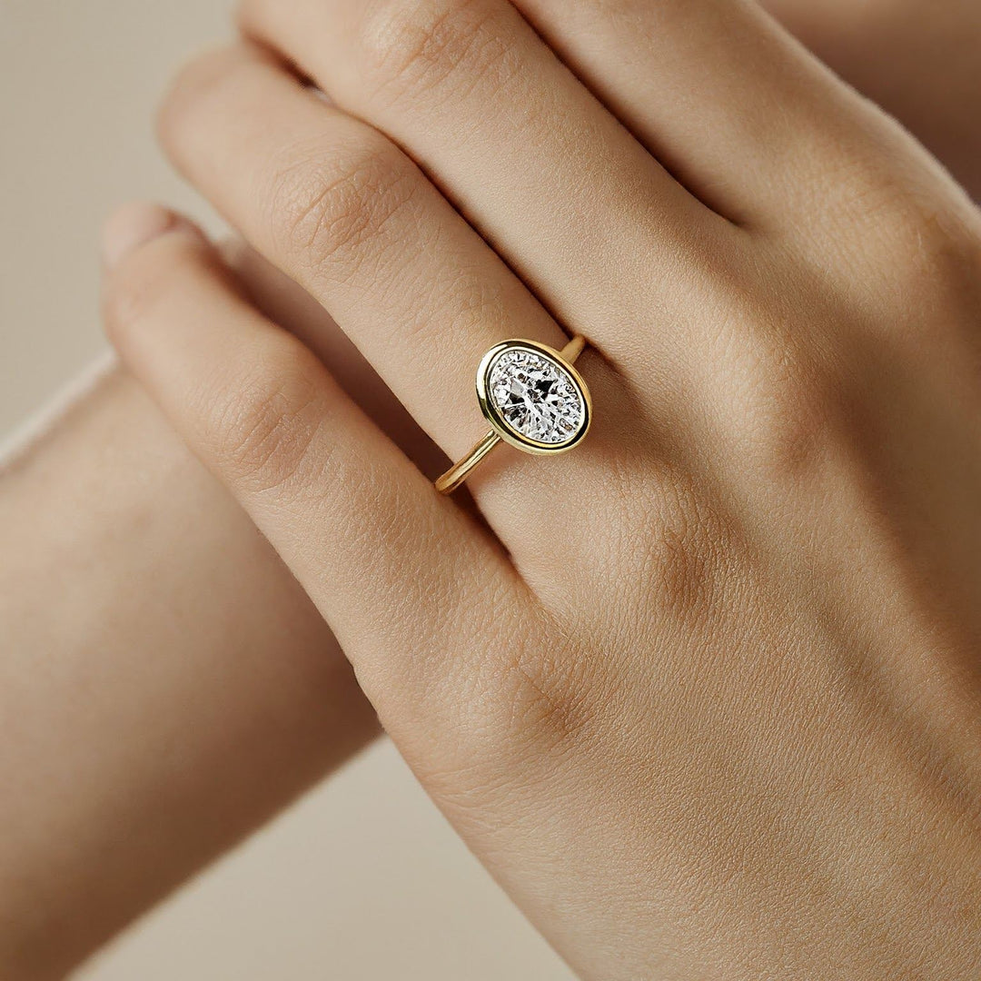 Gold ring with an oval-cut diamond set in a bezel setting worn on a woman's hand.