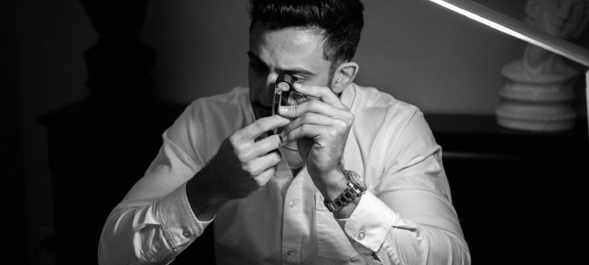 Man in white shirt examining gemstone with a jeweler's loupe under a desk lamp.
