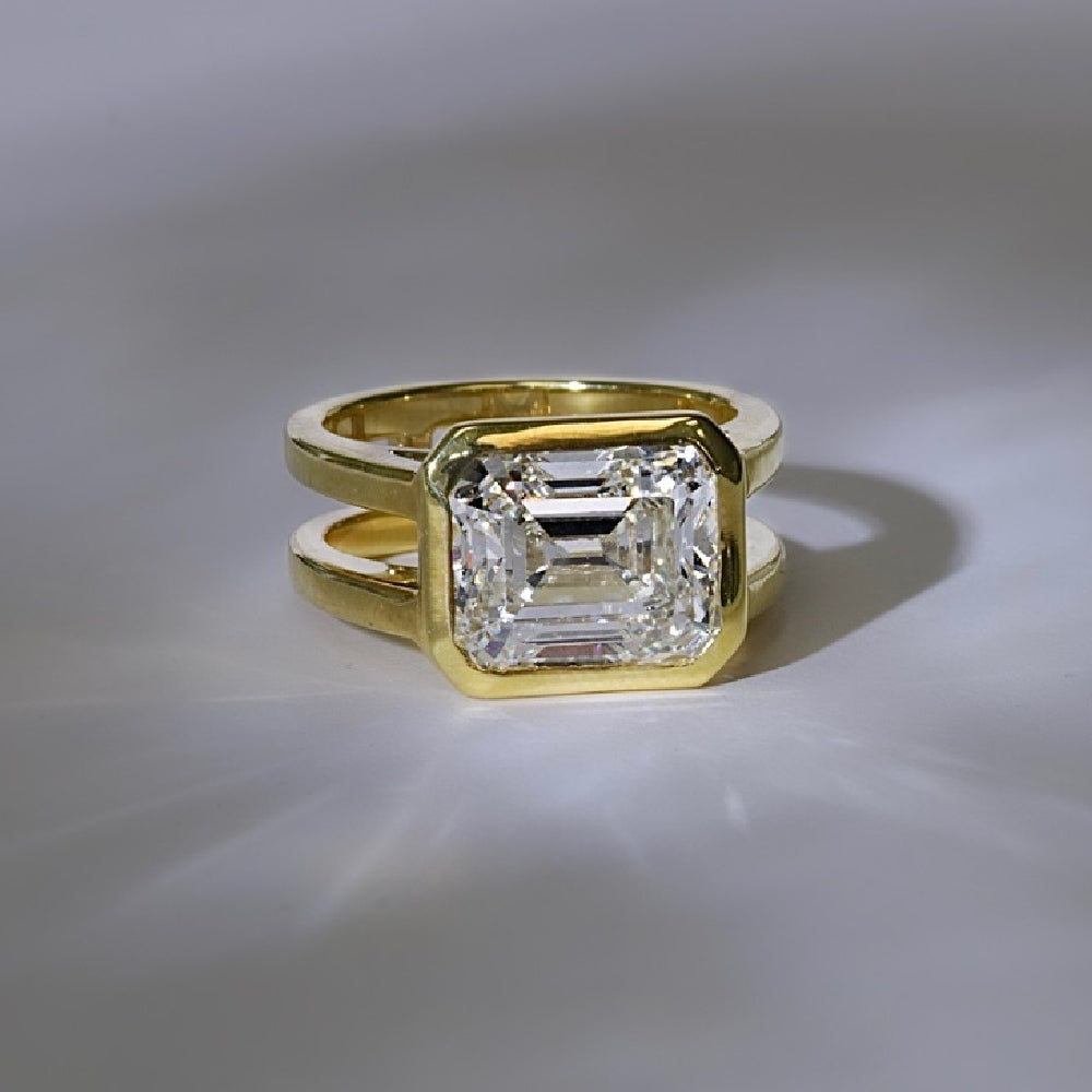 Gold ring with split band featuring a large, emerald-cut diamond in a bezel setting on a light background.