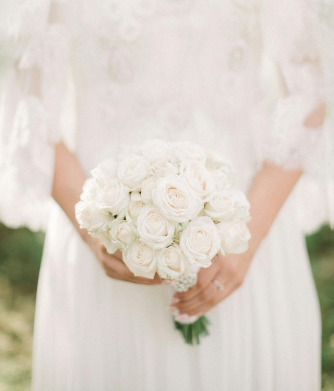 Bride holding a bouquet of white roses, wearing a lace wedding dress in an outdoor setting.