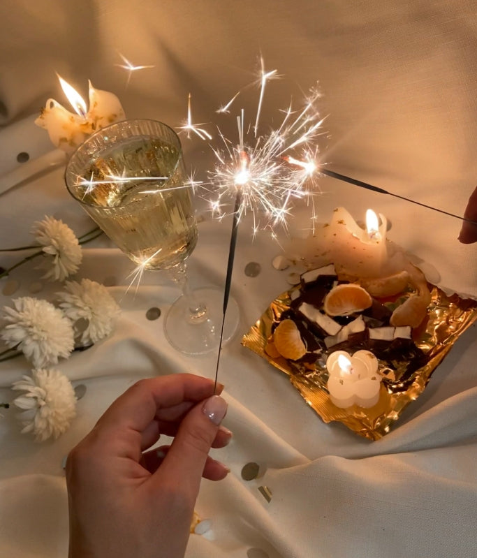 Hands holding sparklers near a glass of champagne, candles, and chocolate with orange slices on gold foil.