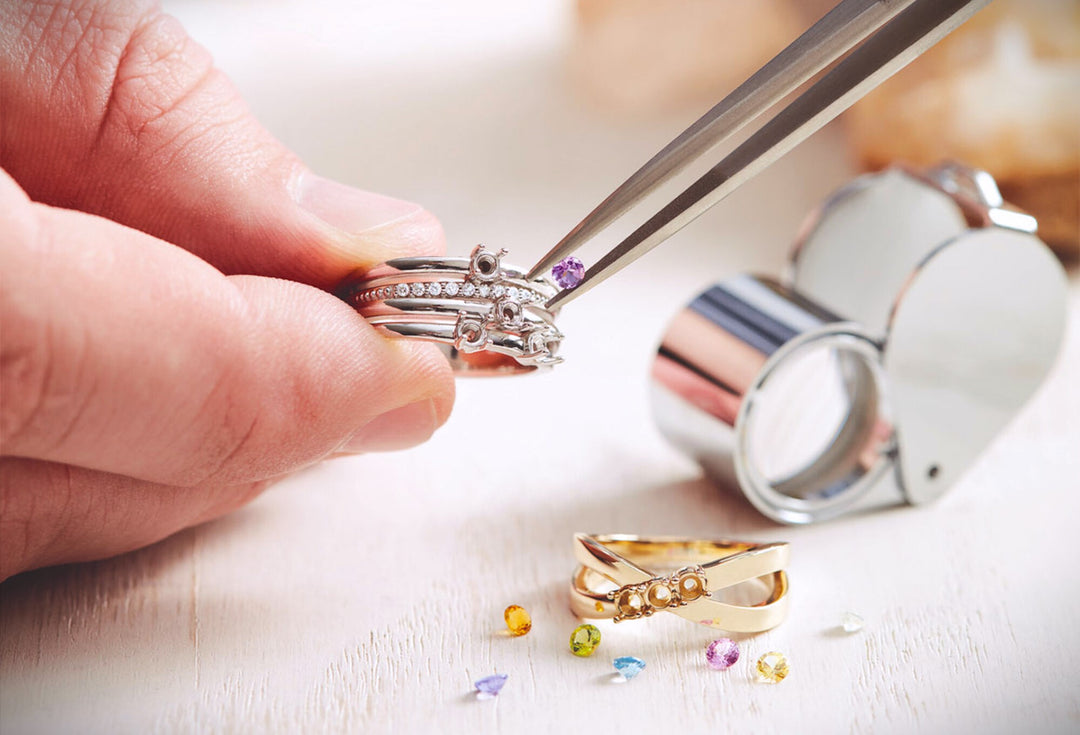 Hand setting a purple gemstone into a silver ring with diamonds, surrounded by loose gems and a jeweler's loupe.