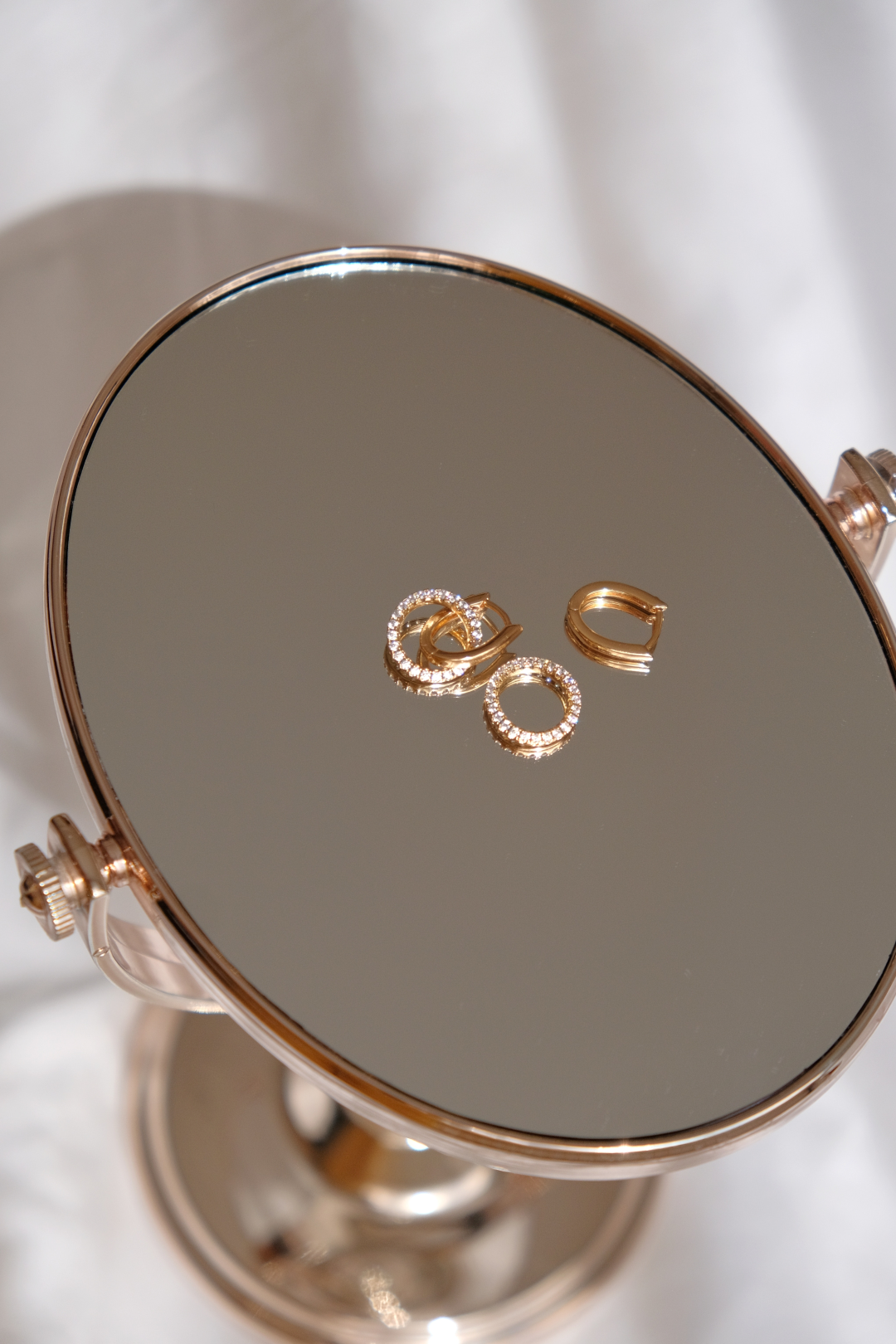 Gold hoop earrings with embedded crystals displayed on a rose gold tabletop mirror.