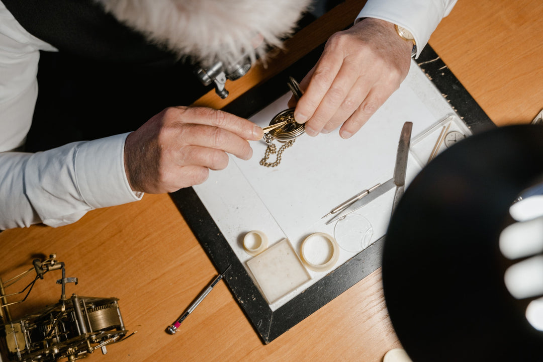 Watchmaker repairing a gold pocket watch under a desk lamp with precision tools on a work mat.