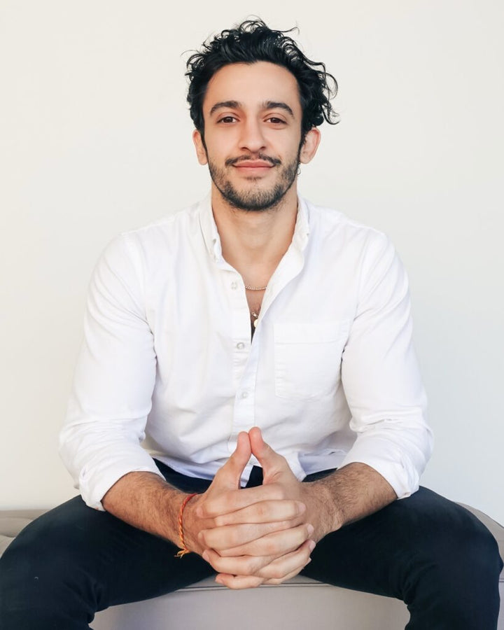 Man in a white button-up shirt and dark pants sitting against a plain white background, hands clasped.