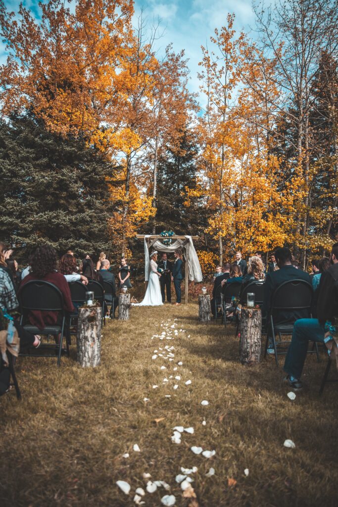Outdoor autumn wedding ceremony with couple at a rustic altar, surrounded by guests and fall-colored trees.