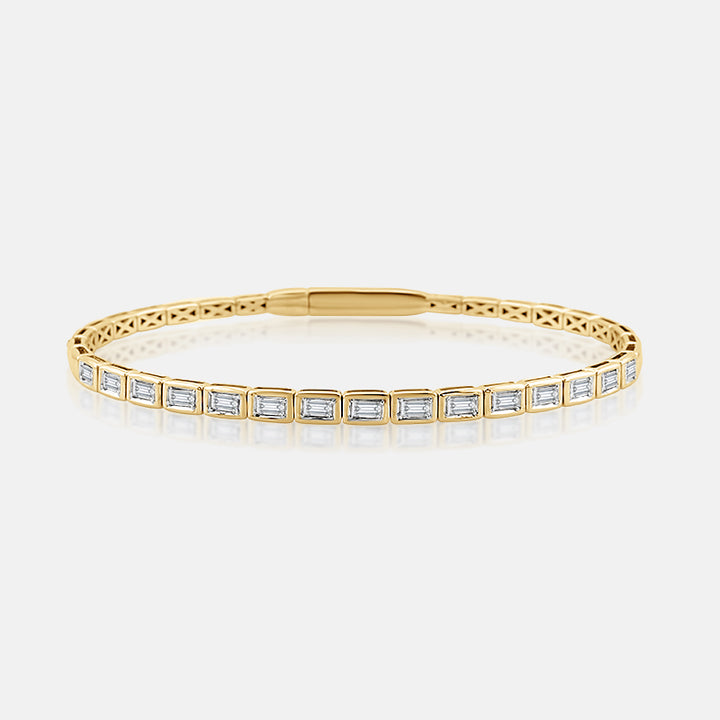 A yellow gold bezel set bracelet featuring emerald cut diamonds, displayed on a clean light gray background with a soft reflection.