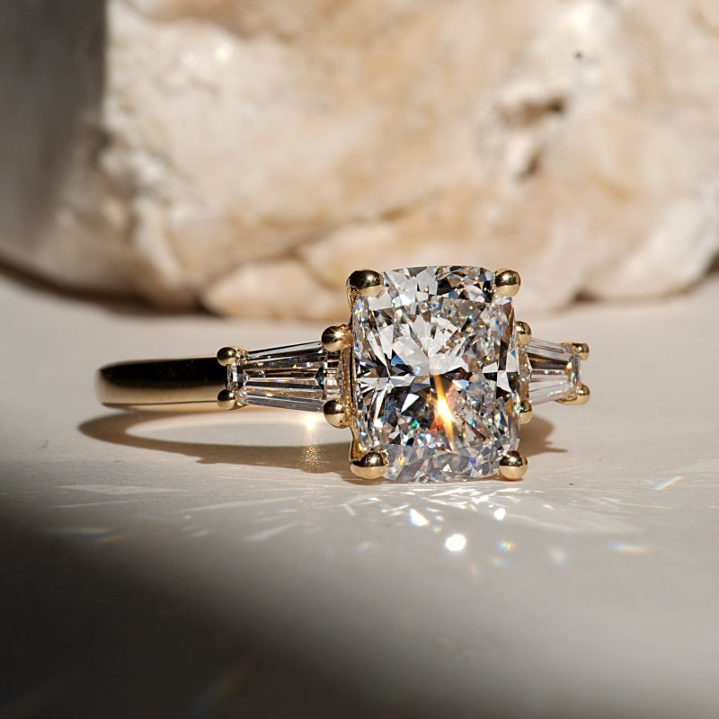 An engagement ring with a large cushion-cut diamond center stone and baguette side stones on a gold band, resting on a light surface with a blurred stone in the background.