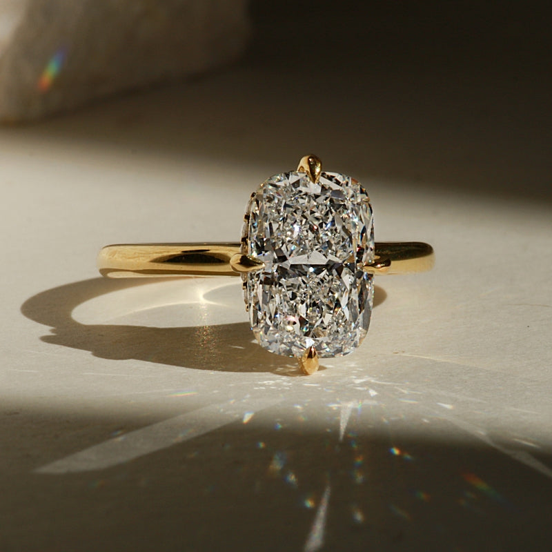 A yellow gold ring with a large elongated  cushion-cut diamond center stone, photographed on a light surface with dramatic sunlight and shadows.