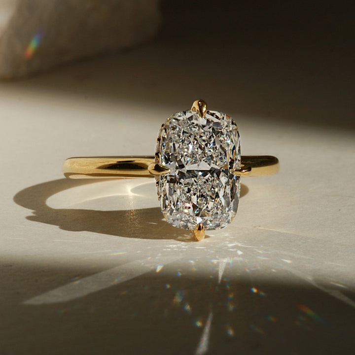 A yellow gold ring with a large elongated  cushion-cut diamond center stone, photographed on a light surface with dramatic sunlight and shadows.
