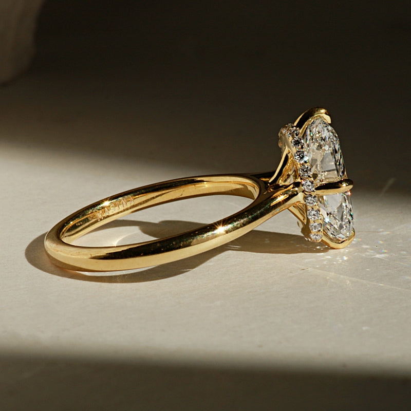 A yellow gold ring with a large elongated cushion cut diamond featuring a hidden halo setting resting on a beige background