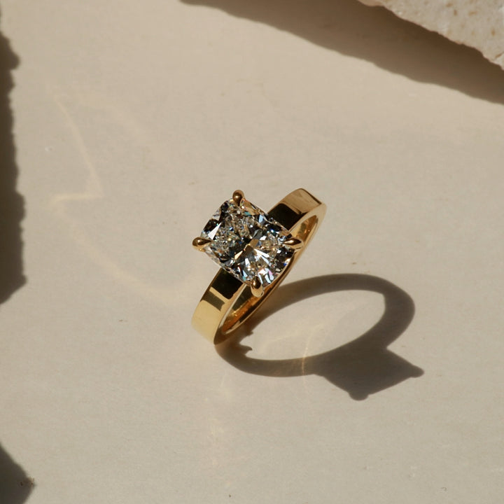 A yellow gold ring with a princess-cut diamond solitaire setting, resting on a textured beige surface with a hard shadow cast to the right.