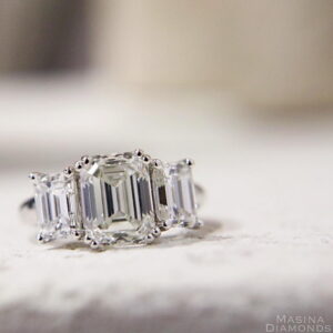 Emerald-cut diamond engagement ring with two side stones set in a platinum band on a light surface.